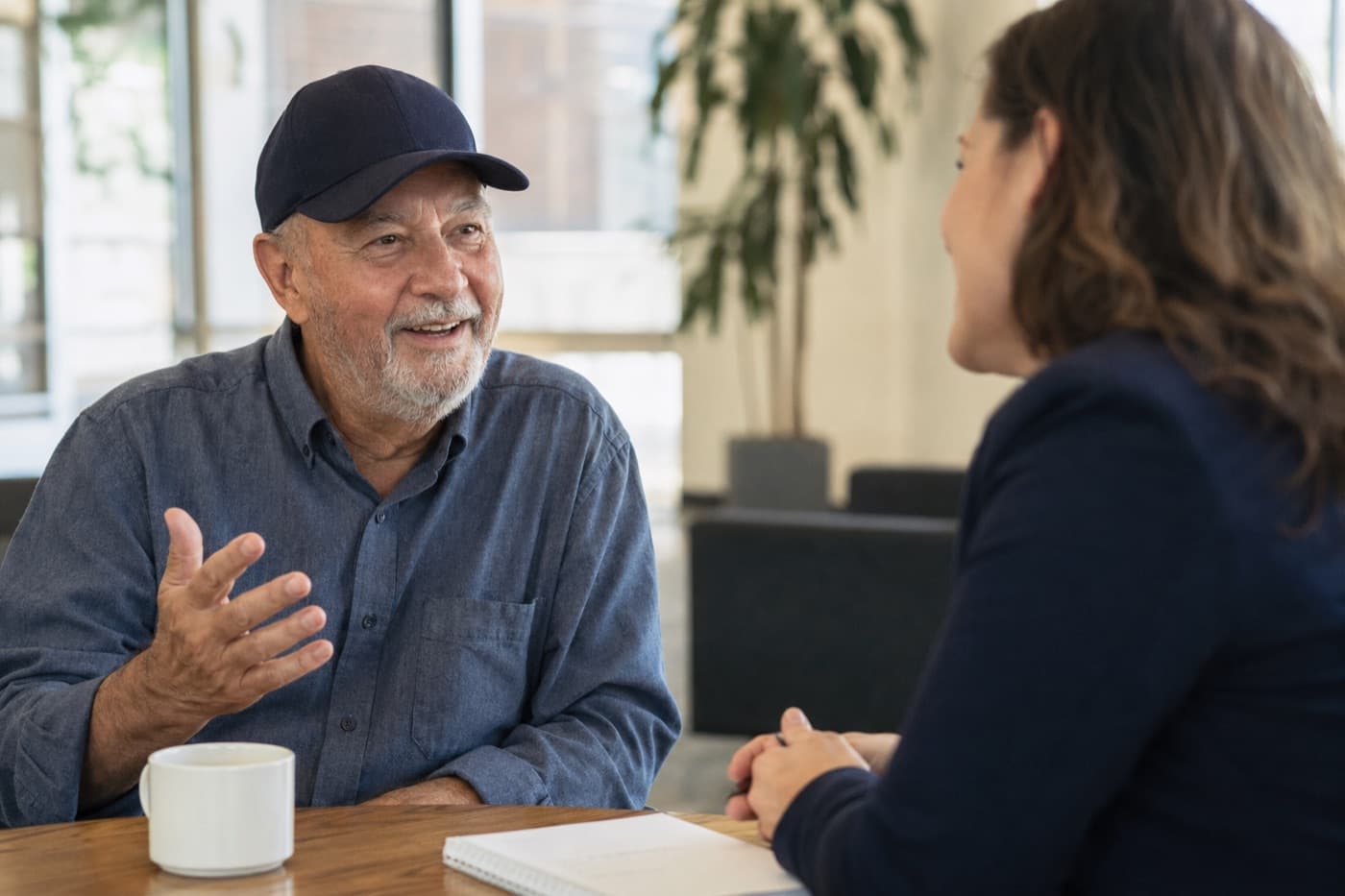A smiling veteran in a navy ball cap talking with an advisor over coffee in a bright office
