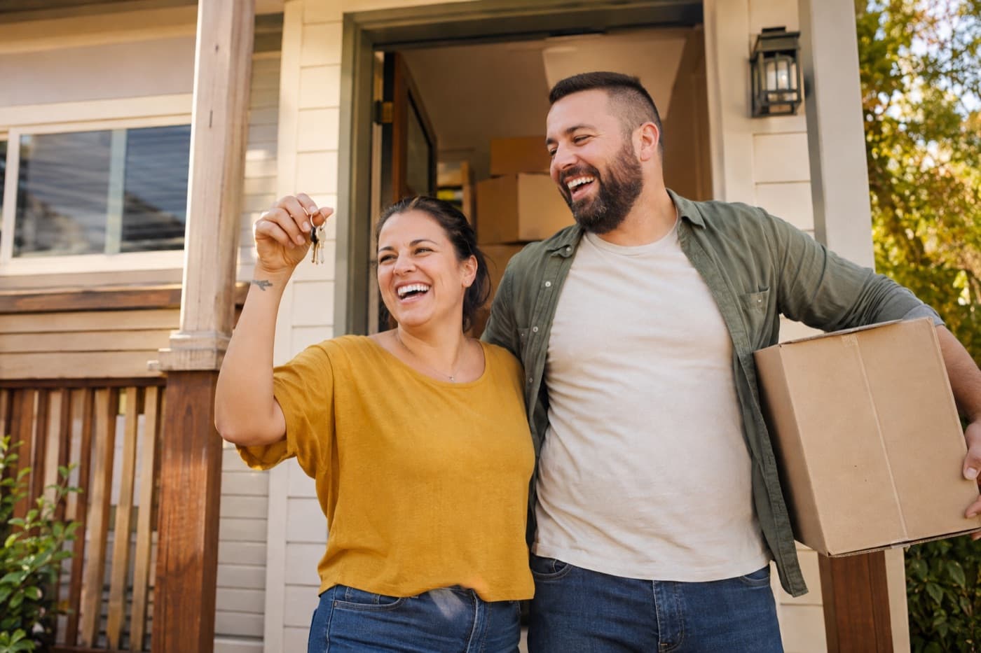 A laughing couple holding up house keys and a moving box on the porch of their new home